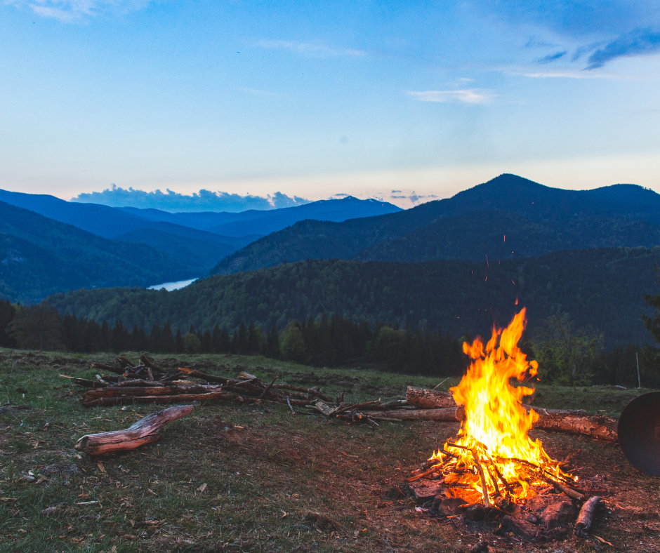 A campfire is in the foreground and mountains in the background with the dawn of the new day behind them