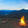 A campfire is in the foreground and mountains in the background with the dawn of the new day behind them